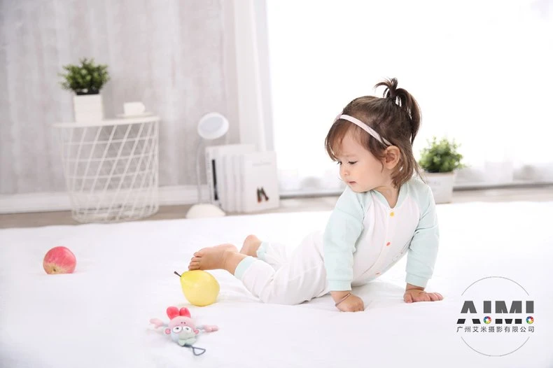 Toddler in a raglan romper prone on a white mat propped on her palms beside toy fruit