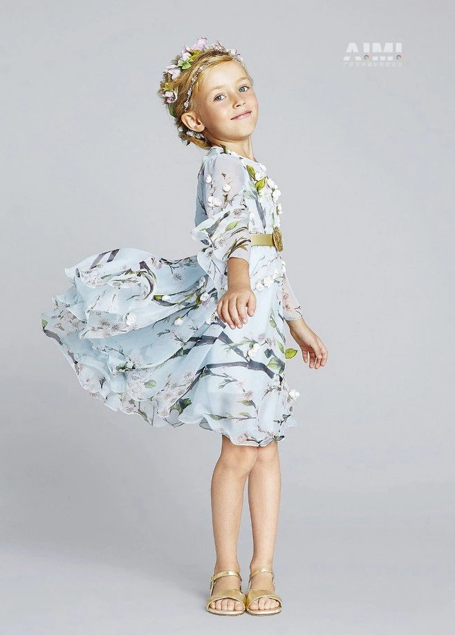 Child model in an ice-blue blossom chiffon dress with gold medallion belt and flower crown, skirt mid-flutter