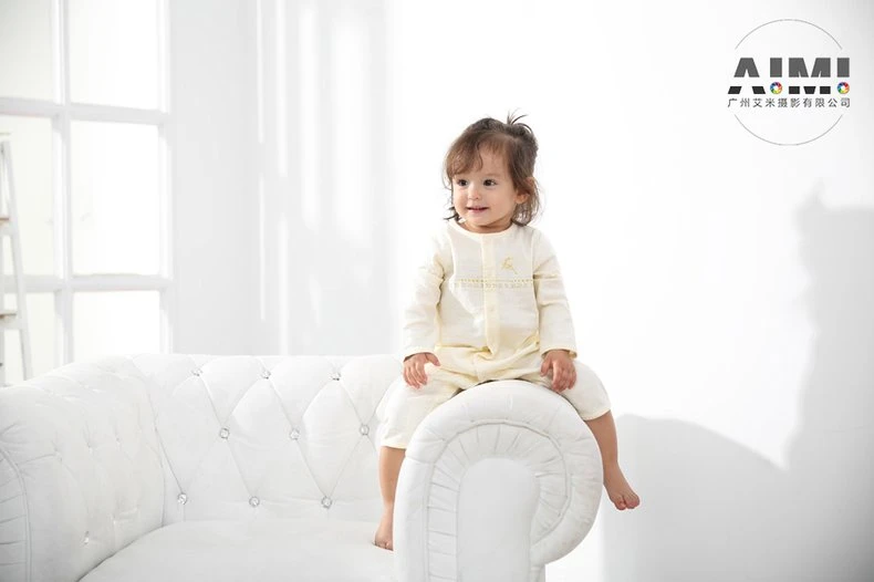 Toddler in a muslin romper straddling the rolled arm of the white sofa, smiling off-camera