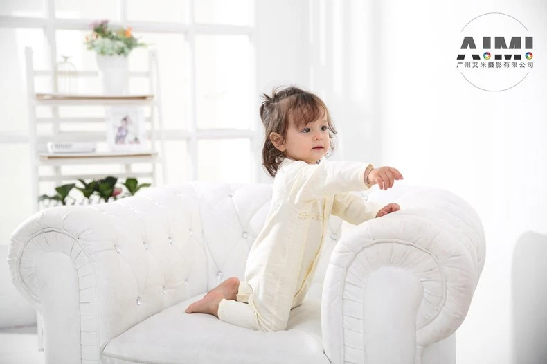 Kneeling toddler in a cream romper in three-quarter back view on the white sofa