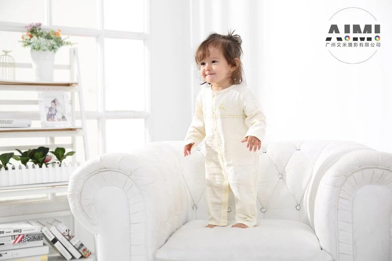 Toddler in a cream romper standing on the chair looking off toward the window