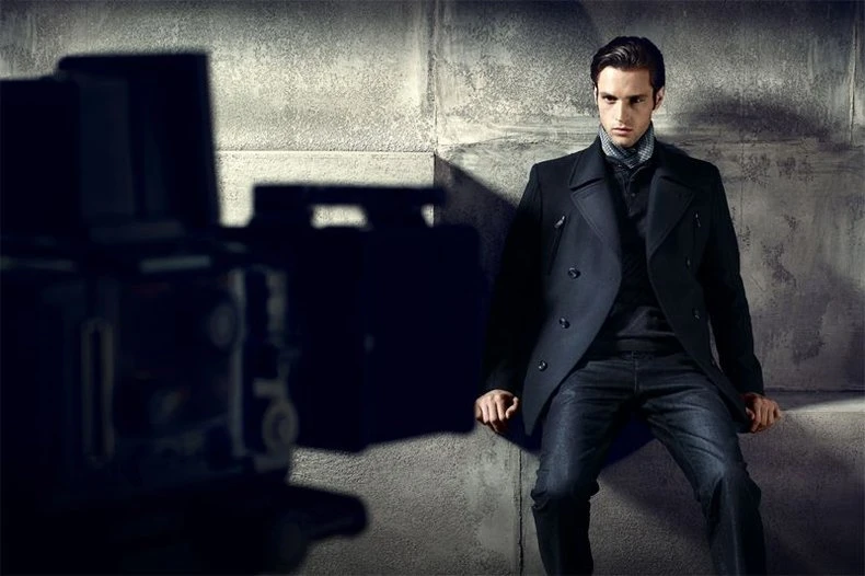 Model in a black double-breasted pea coat seated on a concrete ledge, studio camera blurred in the foreground