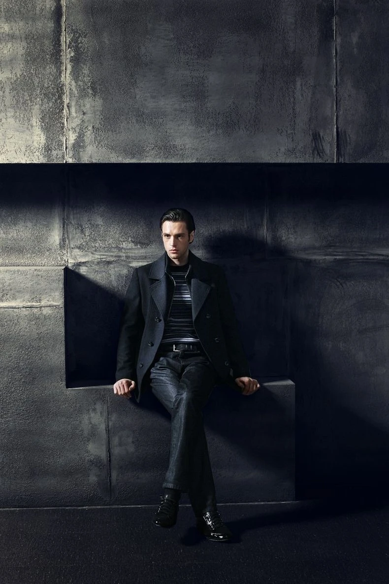 Model in a black wool peacoat seated on a recessed concrete niche with hard shadow play