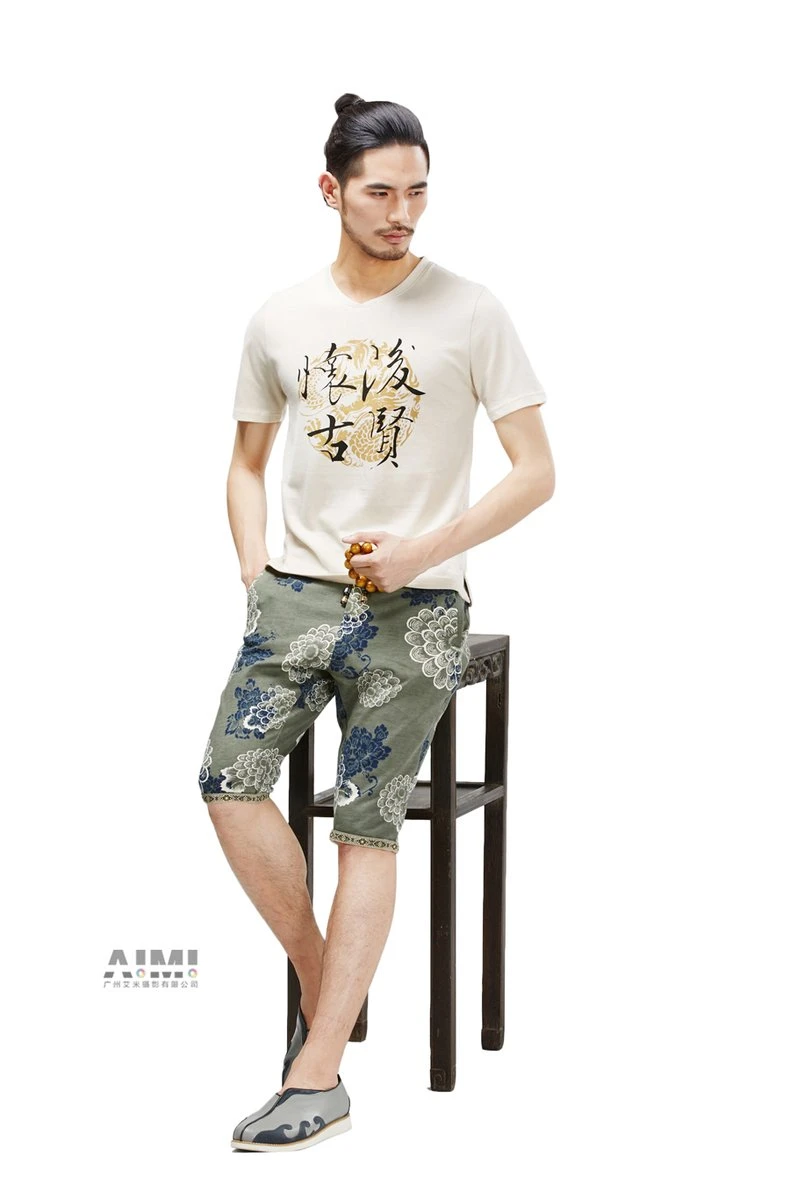 Model in a cream calligraphy tee and sage floral bermuda shorts leaning on a carved hardwood stool