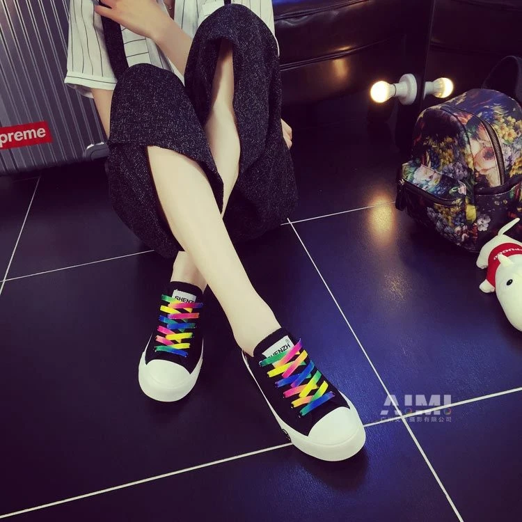 Black canvas sneakers with rainbow gradient laces, crossed-leg on-foot frame on dark glossy tile
